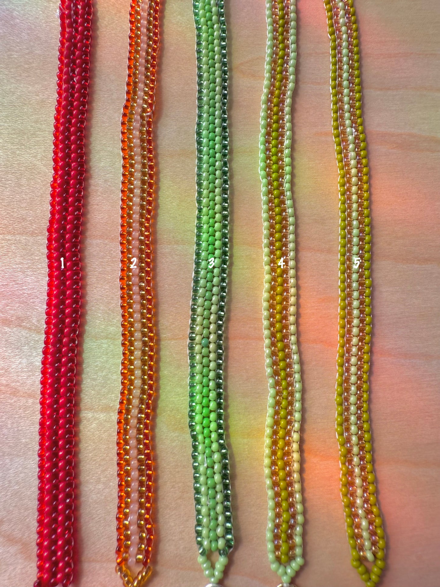 Four beaded necklaces in red, orange, green, and yellow on a textured surface.