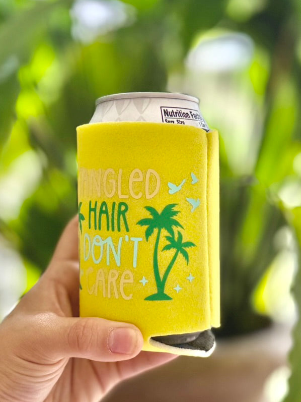 Hand holding a can with a yellow sleeve featuring text and a palm tree design, against a blurred green outdoor background.