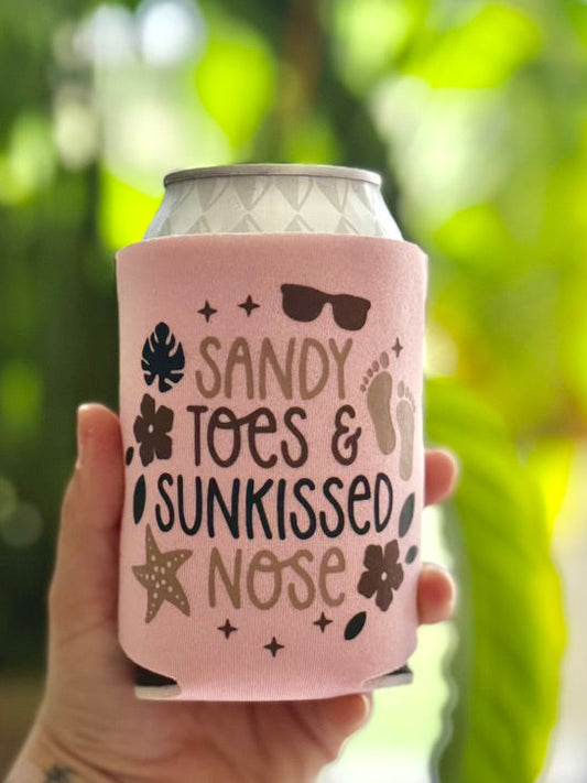 Hand holding a pink can cooler with 'Sandy Toes & Sunkissed Nose' text and beach-themed graphics.