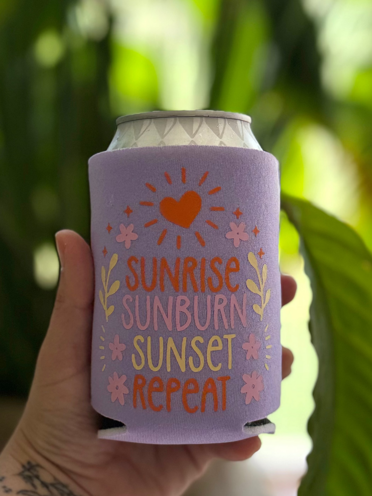 Hand holding a purple can cooler with text and graphics, against a blurred green background