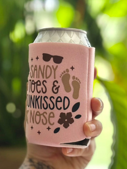 Hand holding a pink can cooler with text and graphics against a blurred green background