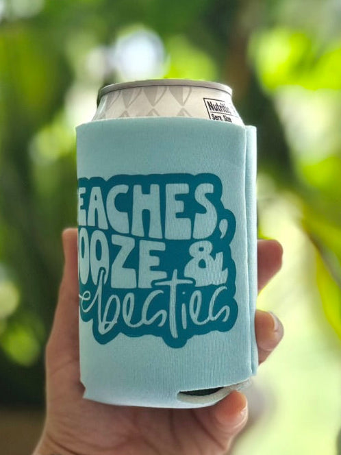 Light blue can cooler with text held by a tattooed hand against a blurred green plant background