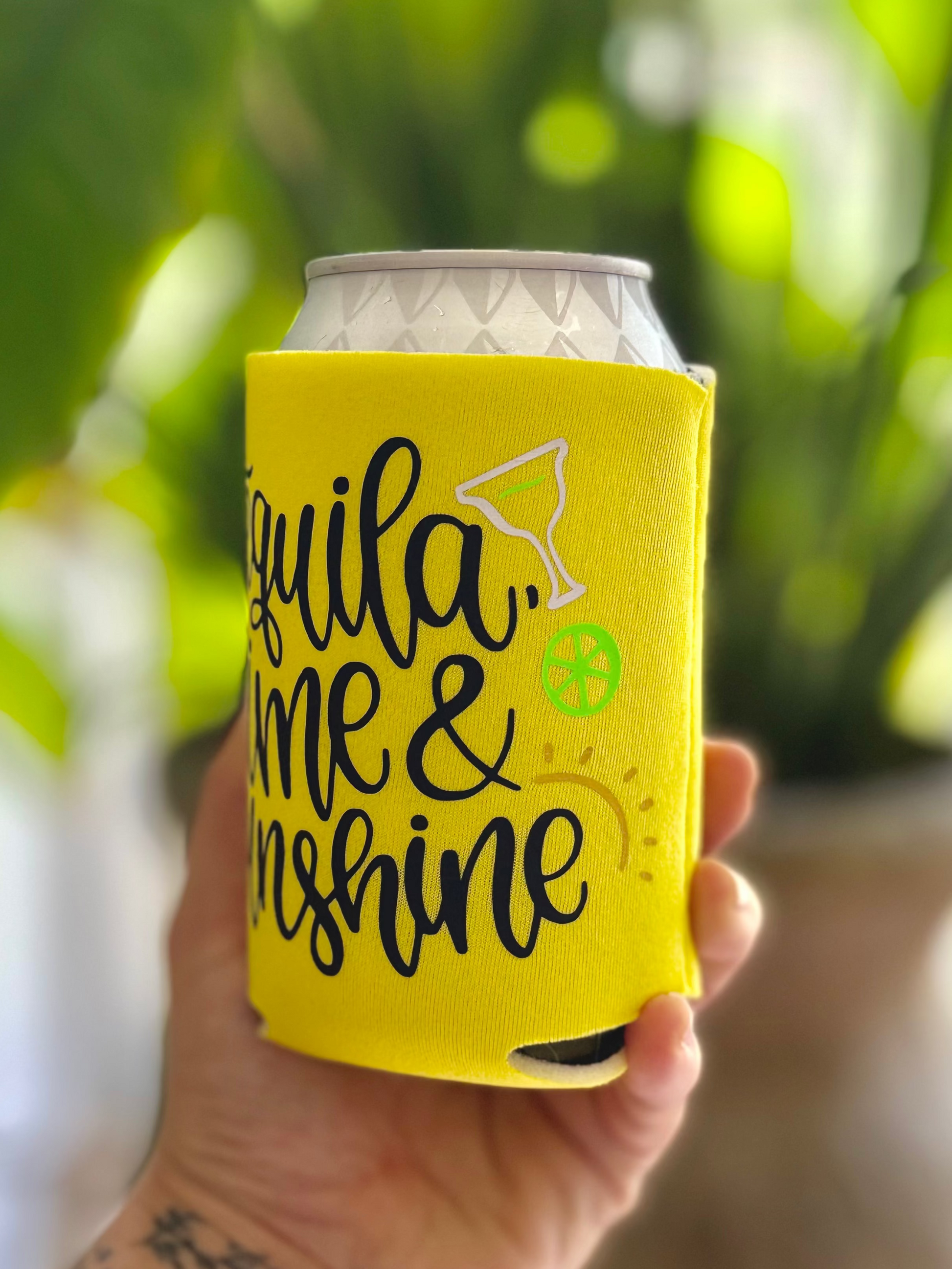 Hand holding a can with a yellow sleeve that says 'Tequila, Me & Sunshine' against a blurred green background.