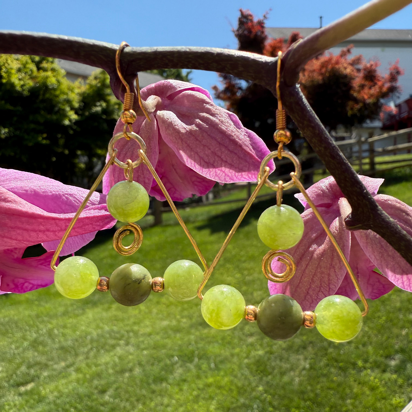Green beaded earrings hanging on a branch with pink flowers and green grass in the background