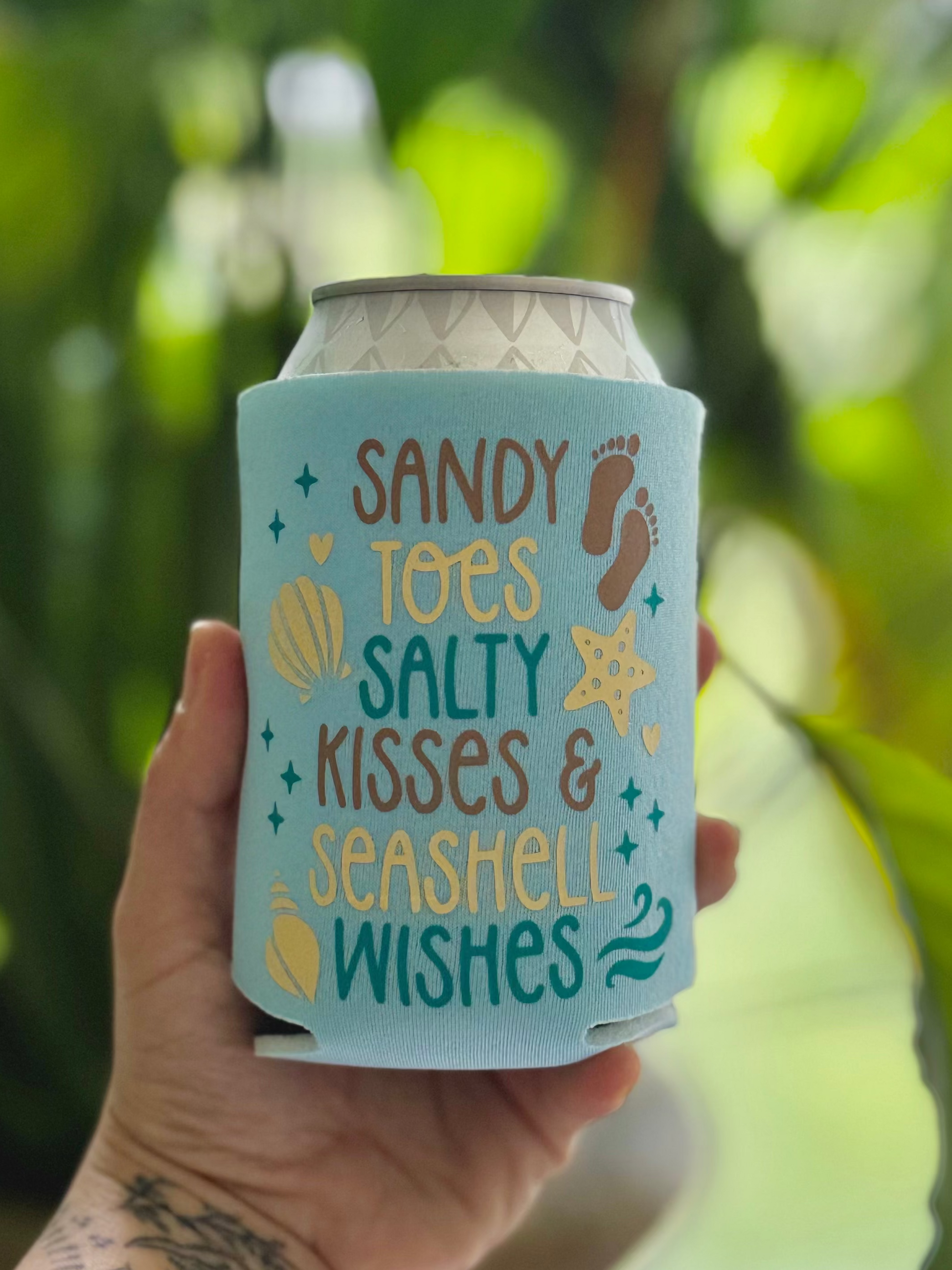 Hand holding a can with a 'Sandy Toes, Salty Kisses & Seashell Wishes' design against a blurred green leafy background.