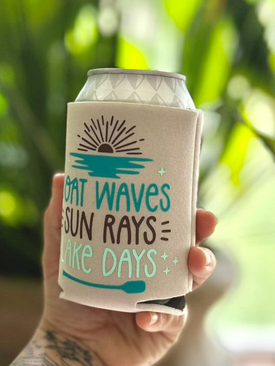 Hand holding a can cooler with 'Waves Sun Rays Lake Days' text against a blurred green background