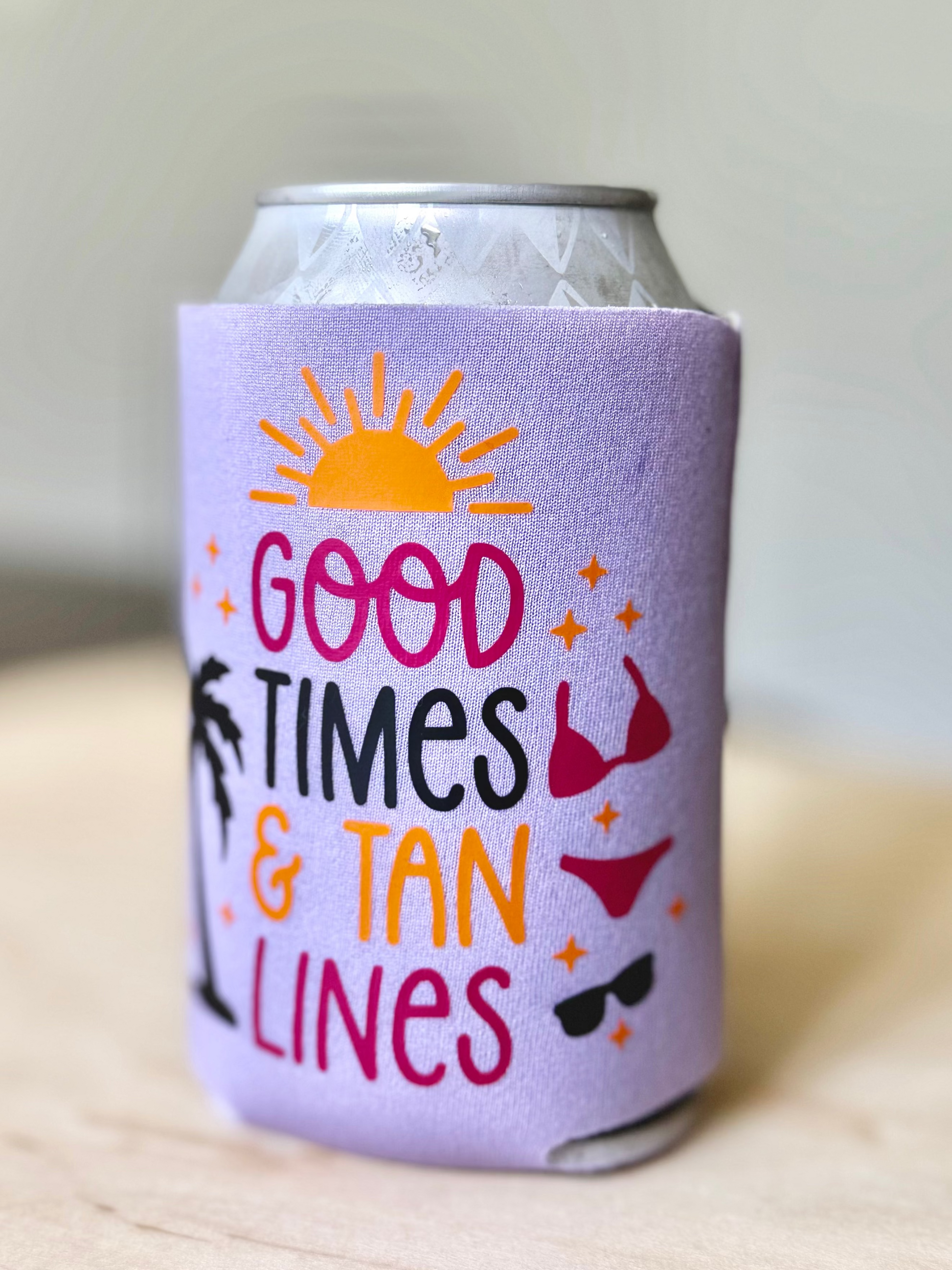 Can cooler with 'Good Times & Tan Lines' text and sun, bikini, and sunglasses graphics on a light background.