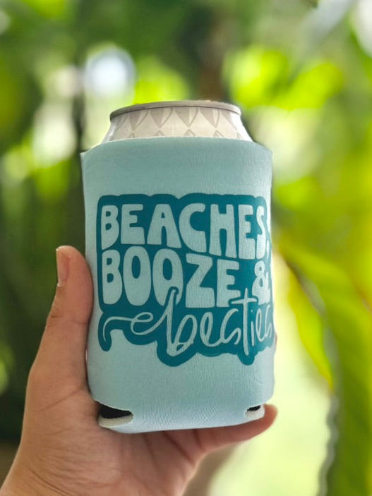 Hand holding a can cooler with 'Beaches, Booze & Boogies' text against a blurred green background