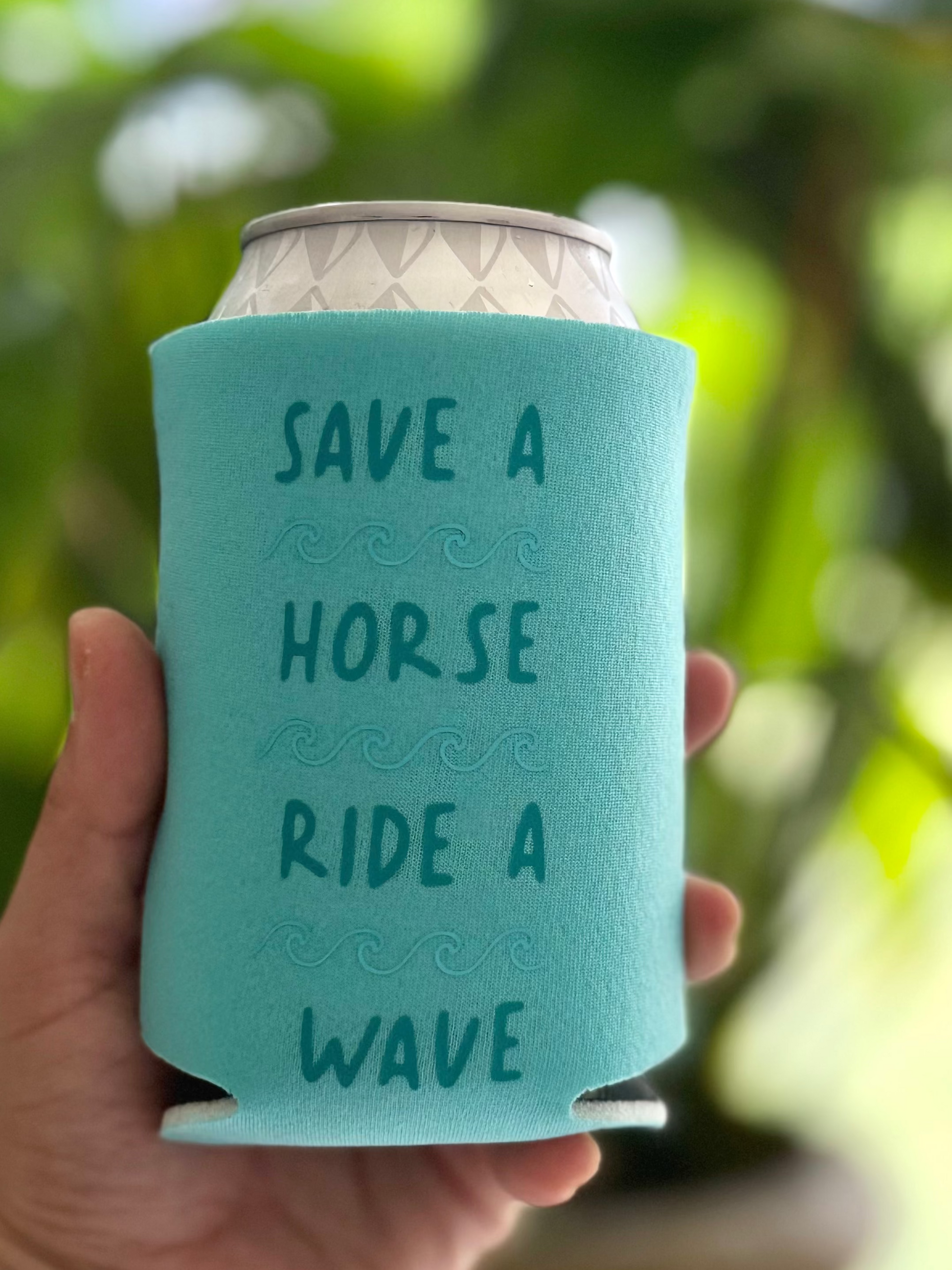 Turquoise can cooler with text held by a hand against a blurred green background
