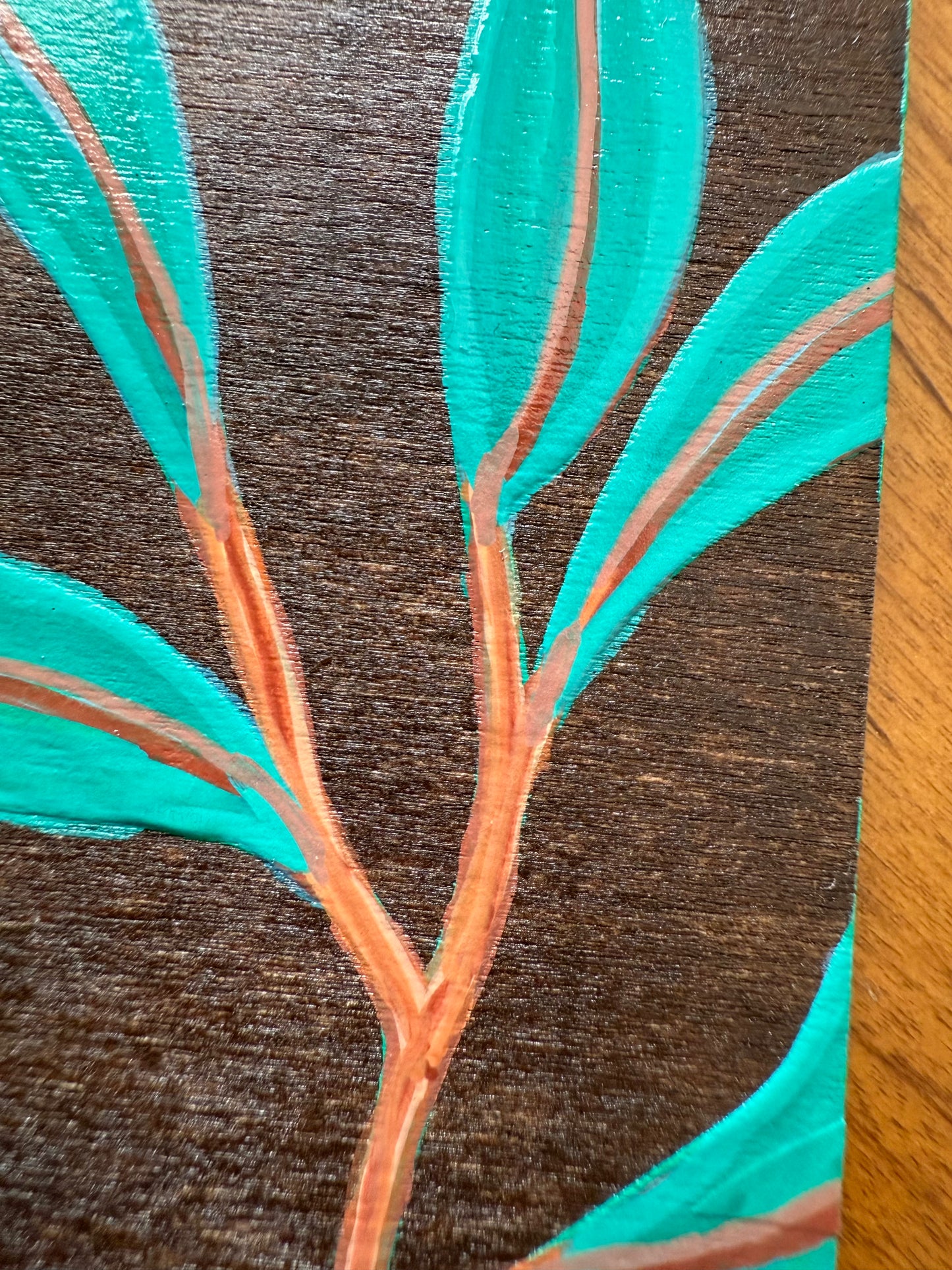 Close-up of a painting of leaves with turquoise leaves and brown stems on a wooden background.