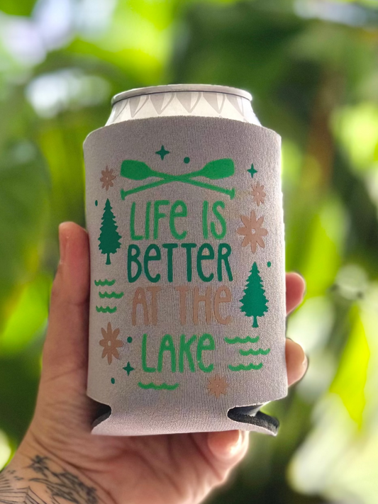 Can cooler with 'Life is Better at the Lake' design held by a tattooed hand against a blurred green leafy background