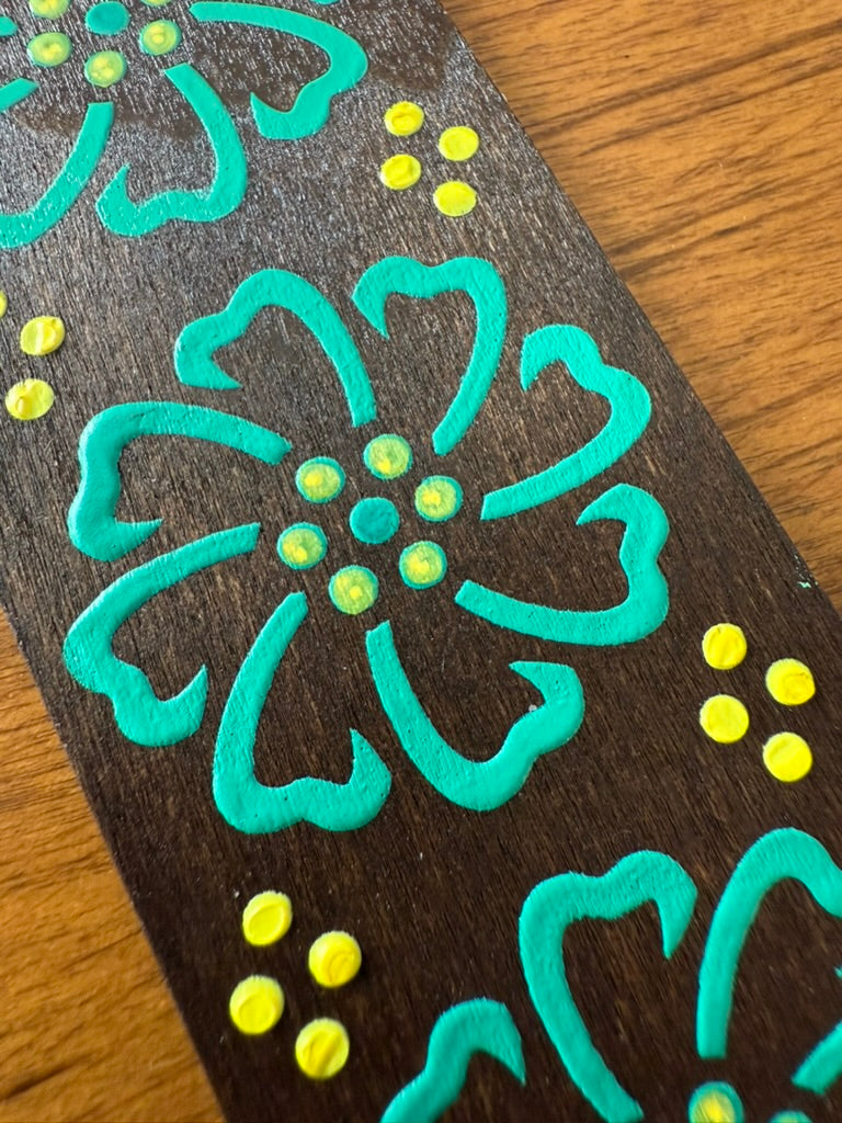 Decorative wooden bookmark with teal floral designs and yellow centers on a wooden surface.