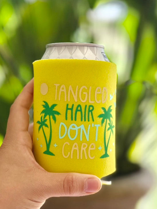 Hand holding a yellow can cooler with 'Tangled Hair Don't Care' text and palm tree designs against a blurred green background.