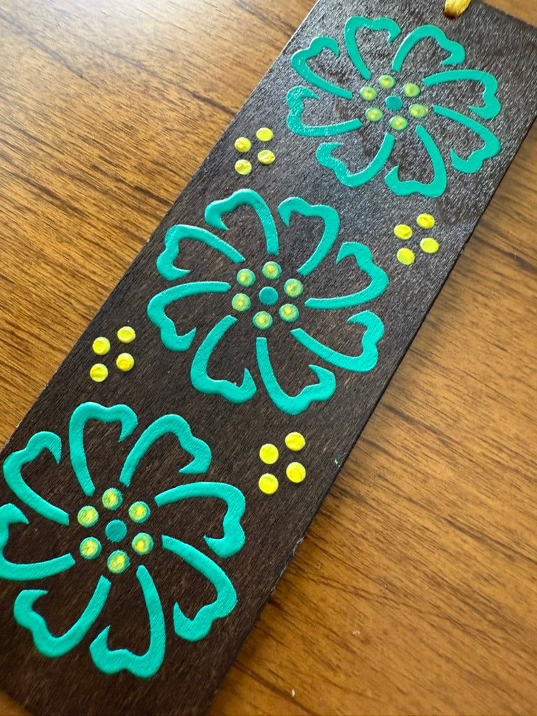 Decorative wooden bookmark with teal floral designs and yellow centers on a wooden surface