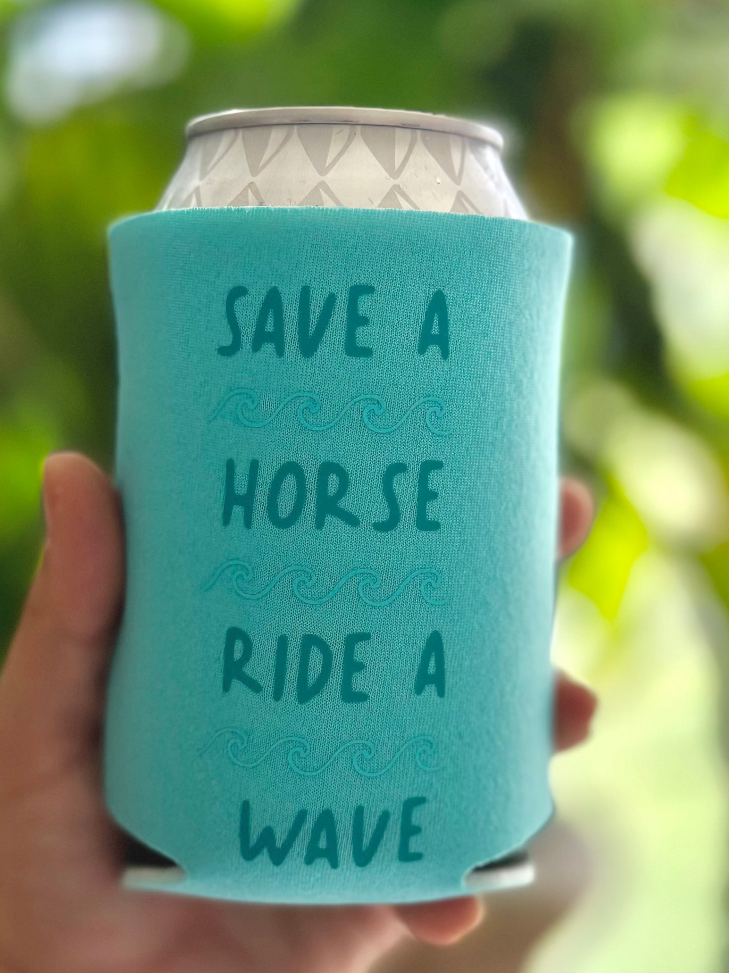 Hand holding a teal can cooler with text against a blurred green background