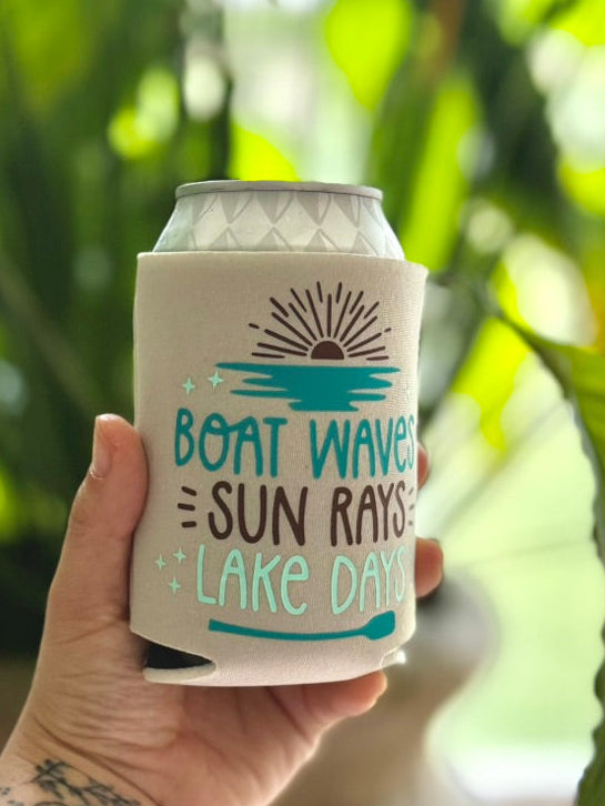 Hand holding a can cooler with 'Boat Waves Sun Rays Lake Days' text against a blurred green background