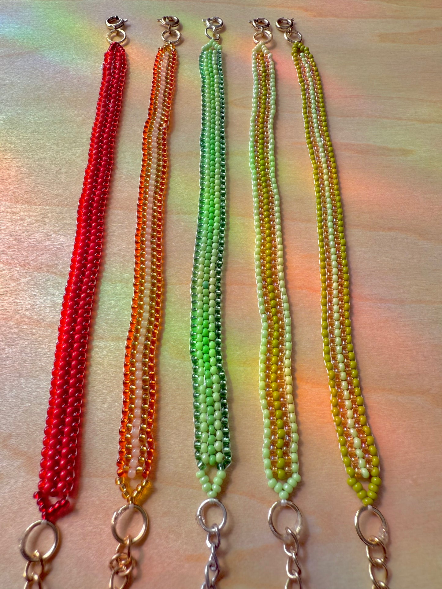 Four beaded necklaces in red, orange, green, and yellow on a textured surface.