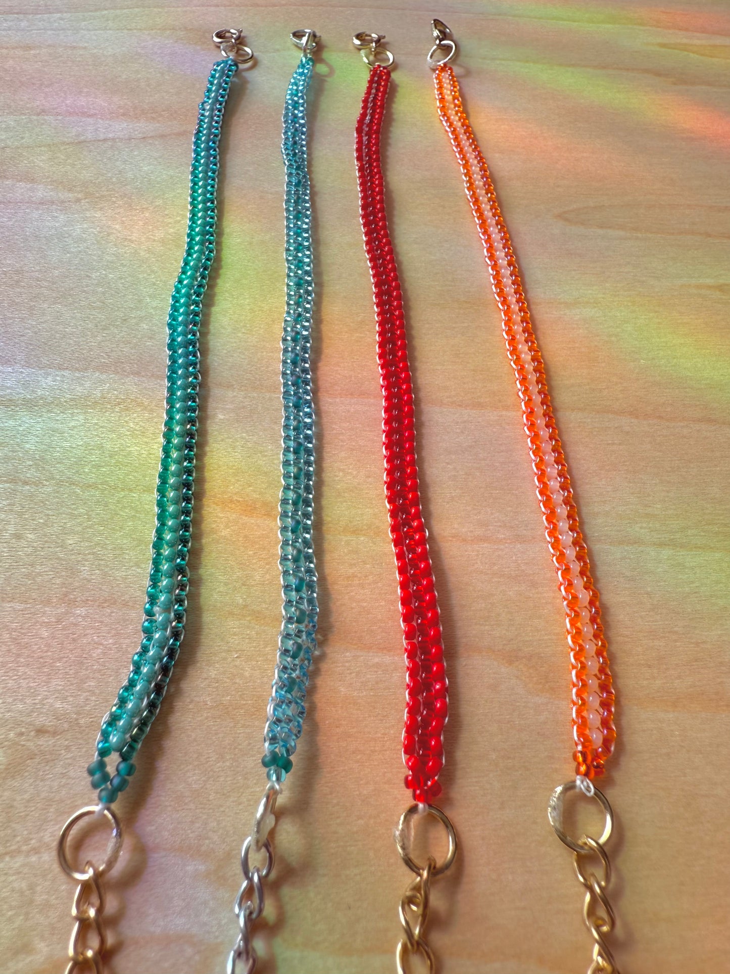 Four beaded necklaces in green, blue, red, and orange on a wooden surface.
