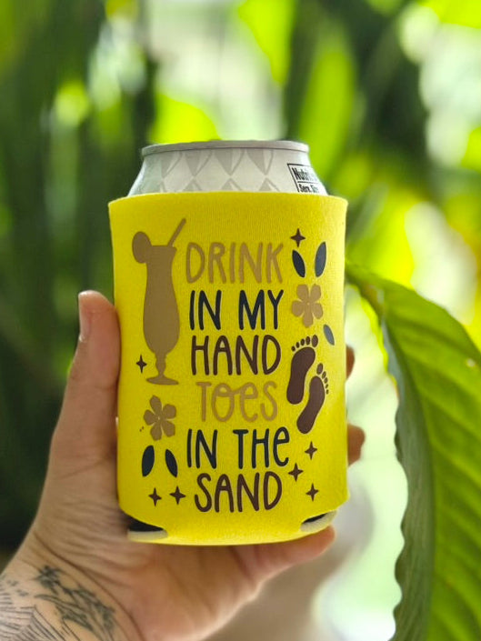 Yellow can cooler with beach-themed design held by a person with tattoos, against a blurred green leafy background.