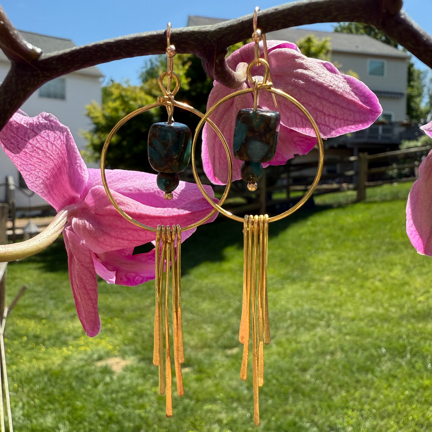 Gold hoop earrings with dark beads hanging from pink flowers against a grassy outdoor background.