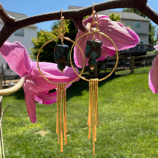 Gold hoop earrings with dark beads hanging from pink flowers against a grassy outdoor background.