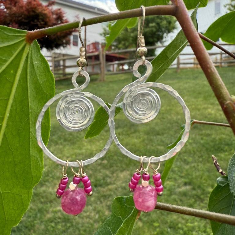 Silver hoop earrings with pink gemstones hanging on a branch with green leaves.