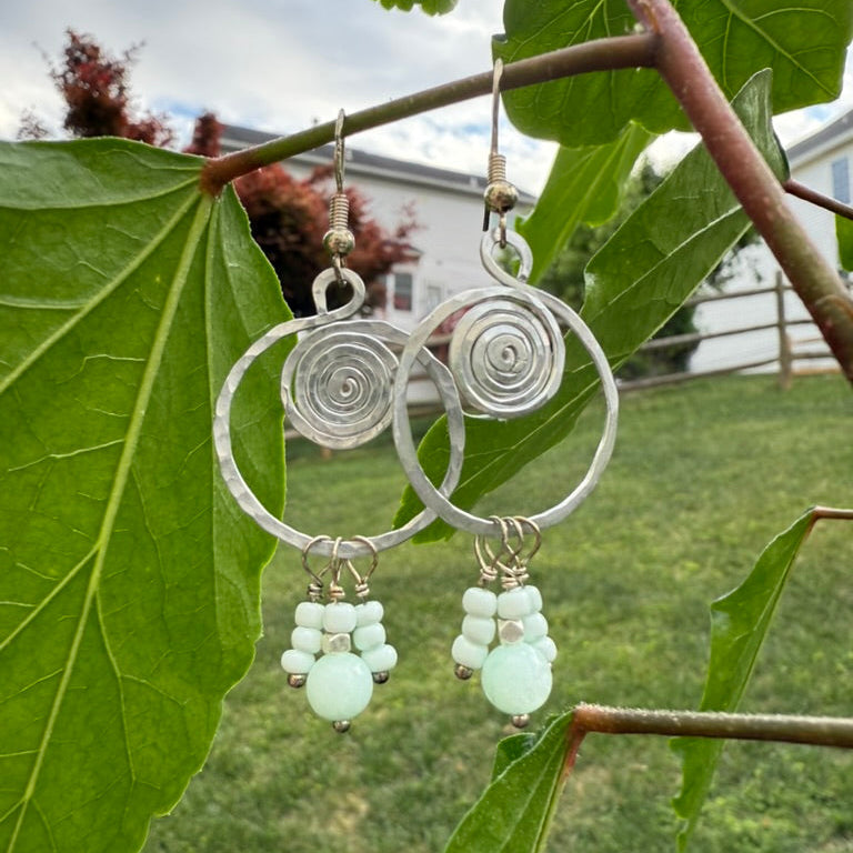 Silver hoop earrings with green beads hanging on a branch with leaves.