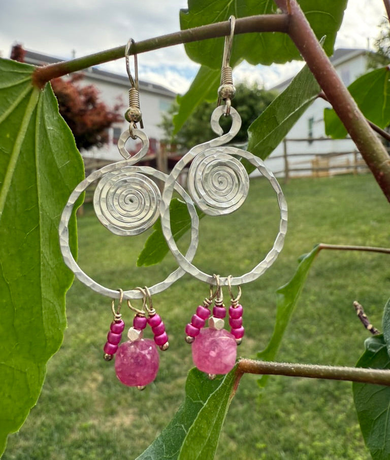 Silver hoop earrings with pink beads hanging on a branch with leaves.