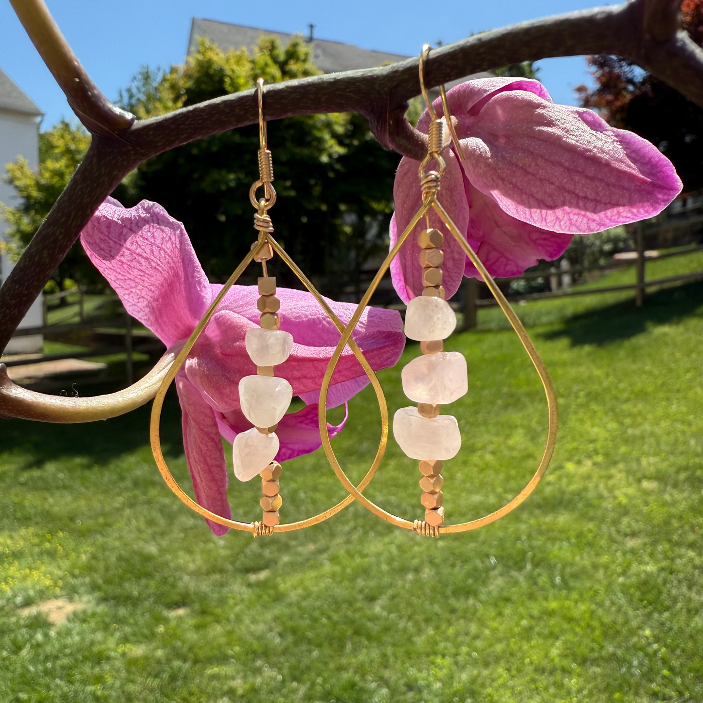 Gold hoop earrings with white beads hanging on pink flowers outdoors.
