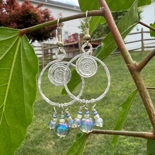 Pair of silver spiral earrings with beads hanging on a branch with green leaves.