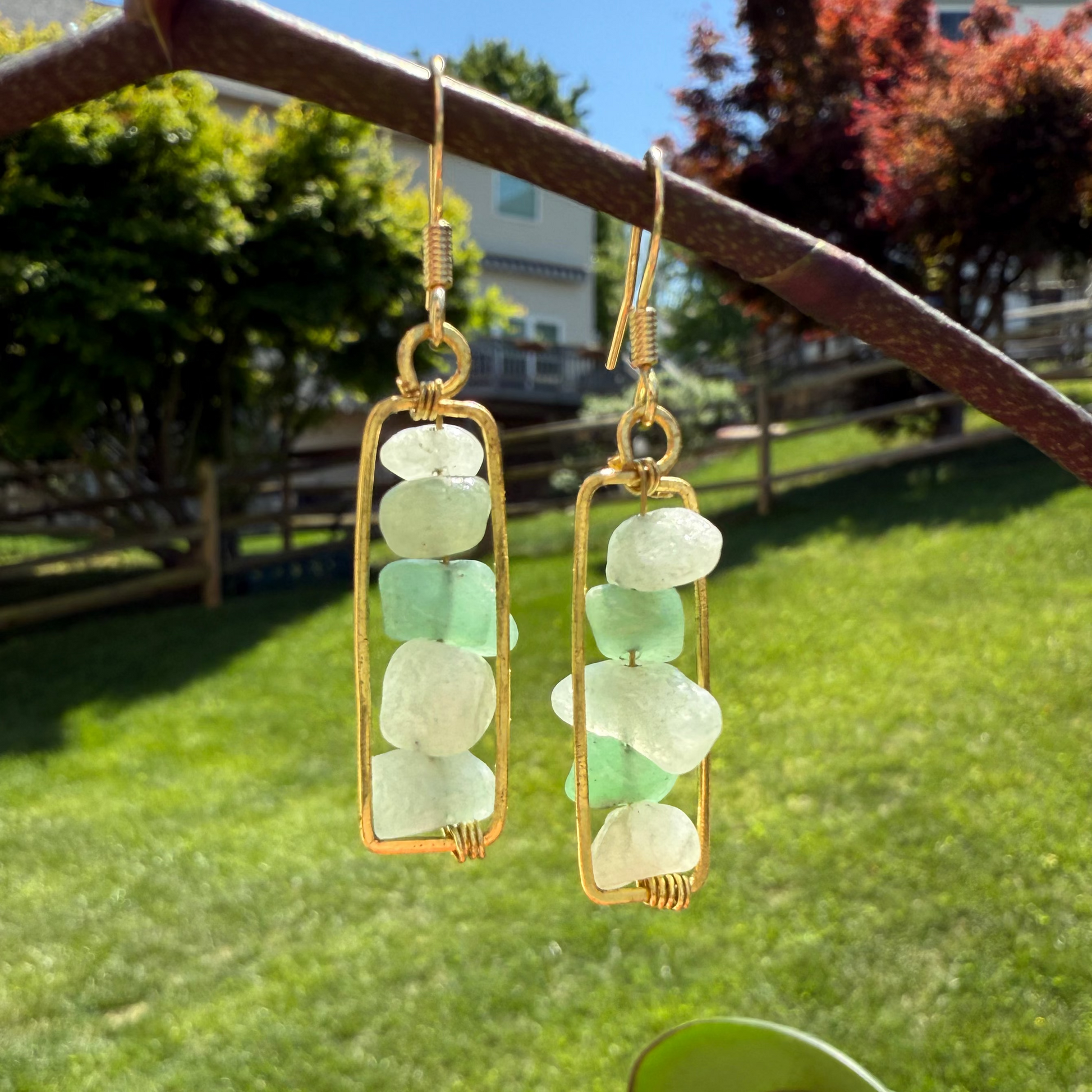 Gold-framed green beaded earrings hanging on a metal bar with a grassy area and trees in the background.
