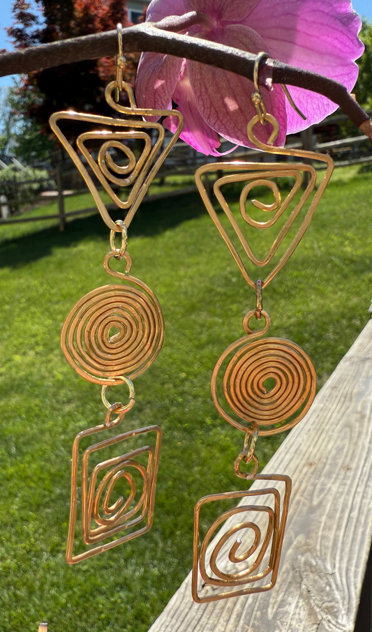 Gold wire wind chime with geometric designs hanging on a wooden railing outdoors.