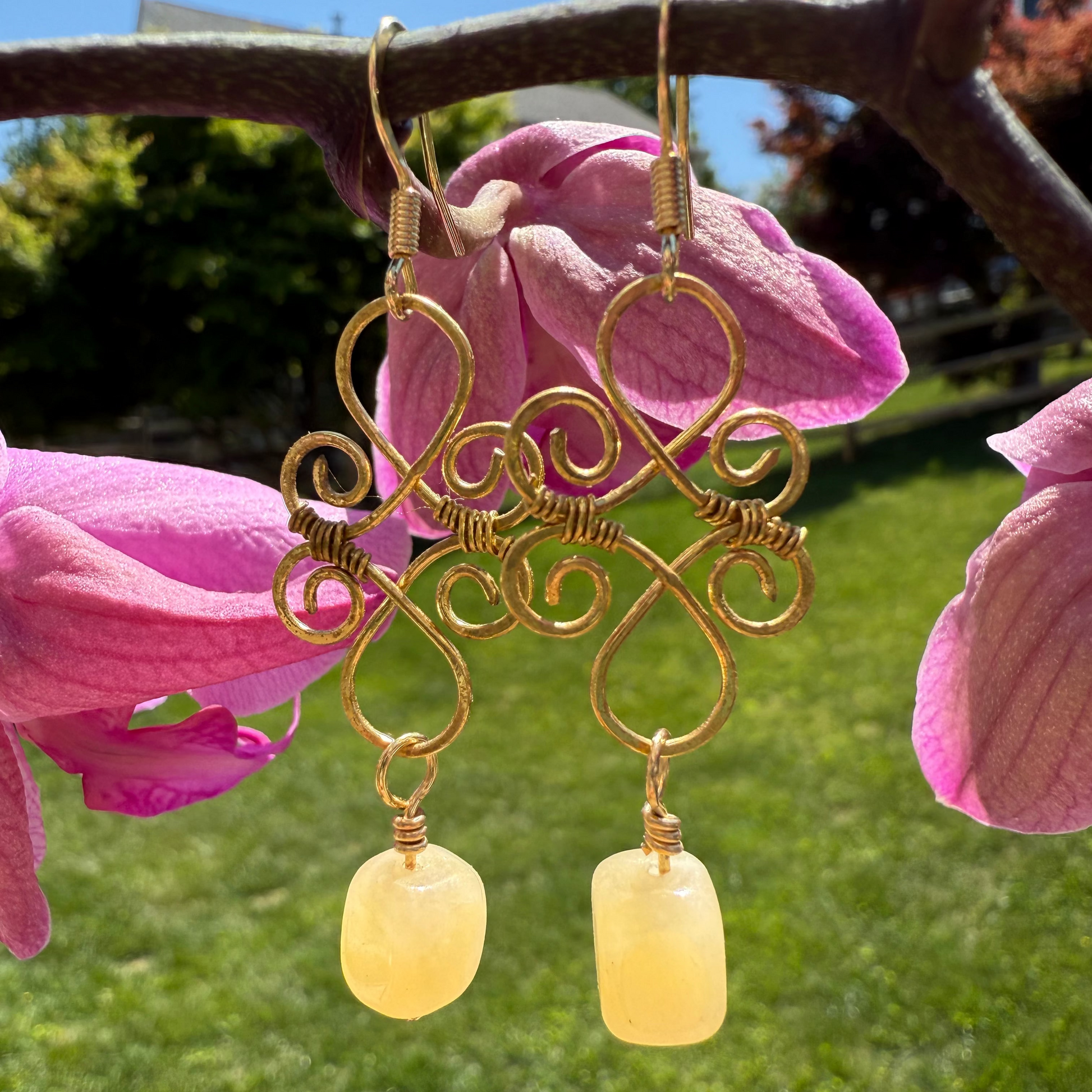 Gold earrings with yellow beads hanging from pink flowers outdoors.