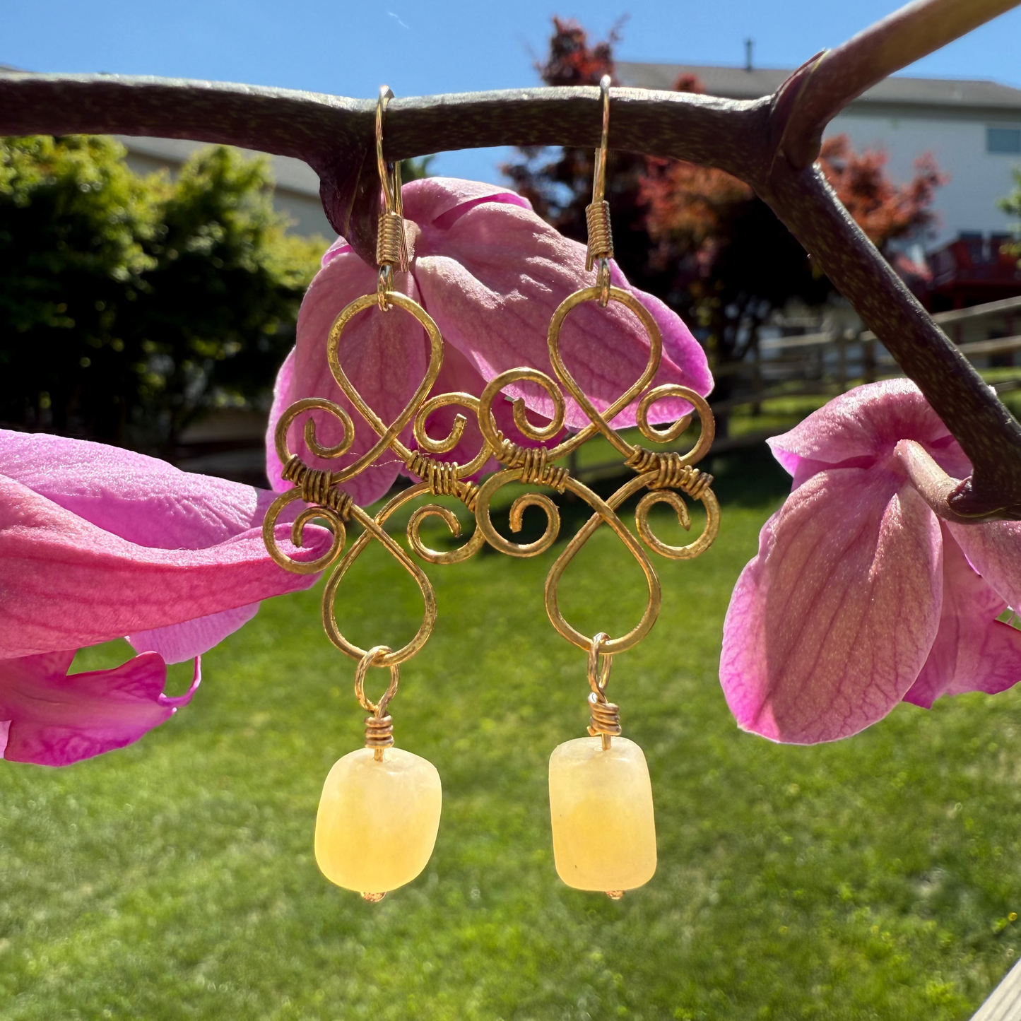 Gold earrings with yellow beads hanging from pink flowers outdoors.